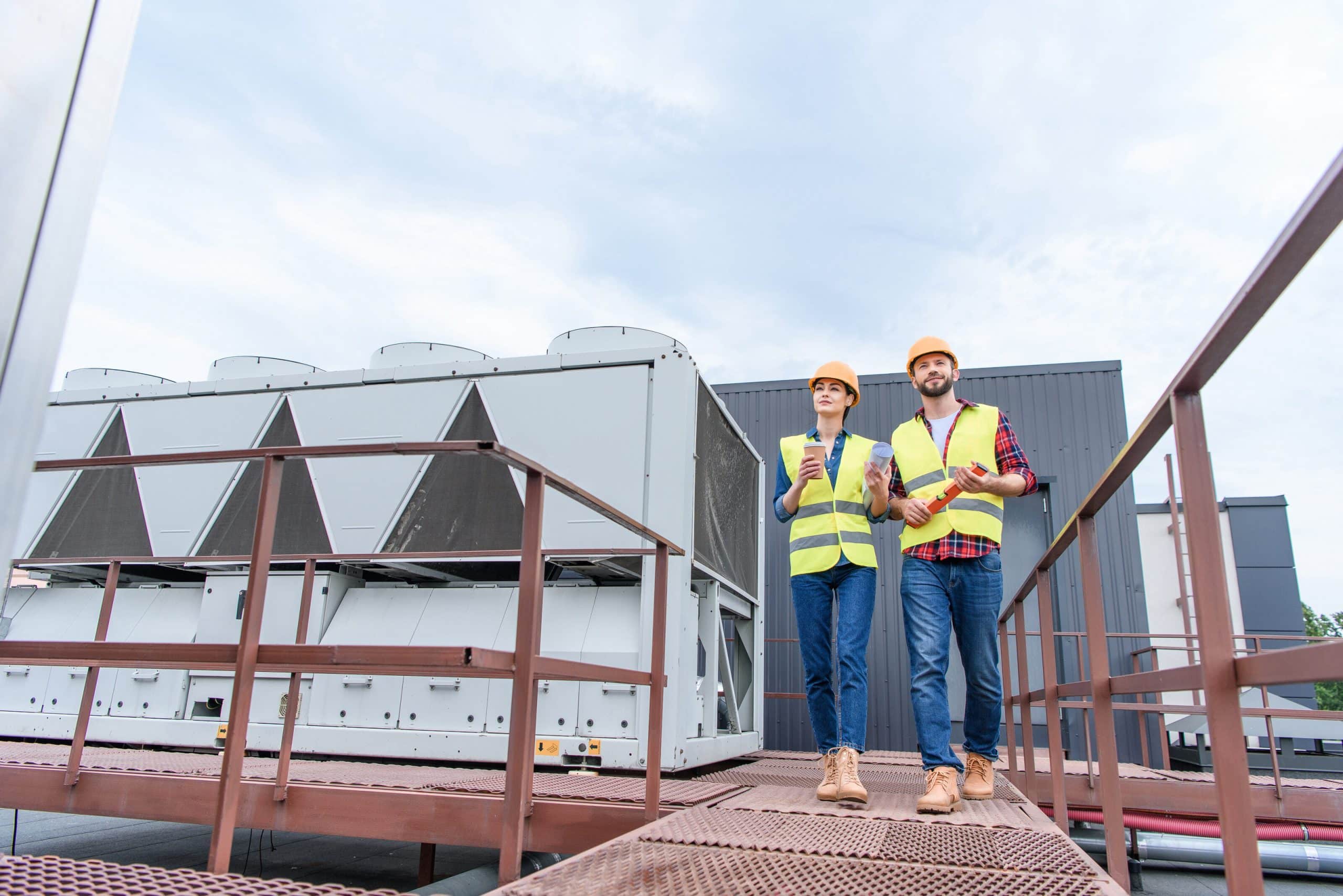 Two engineers in hard hats and high-visibility vests standing beside a large rooftop HVAC system.