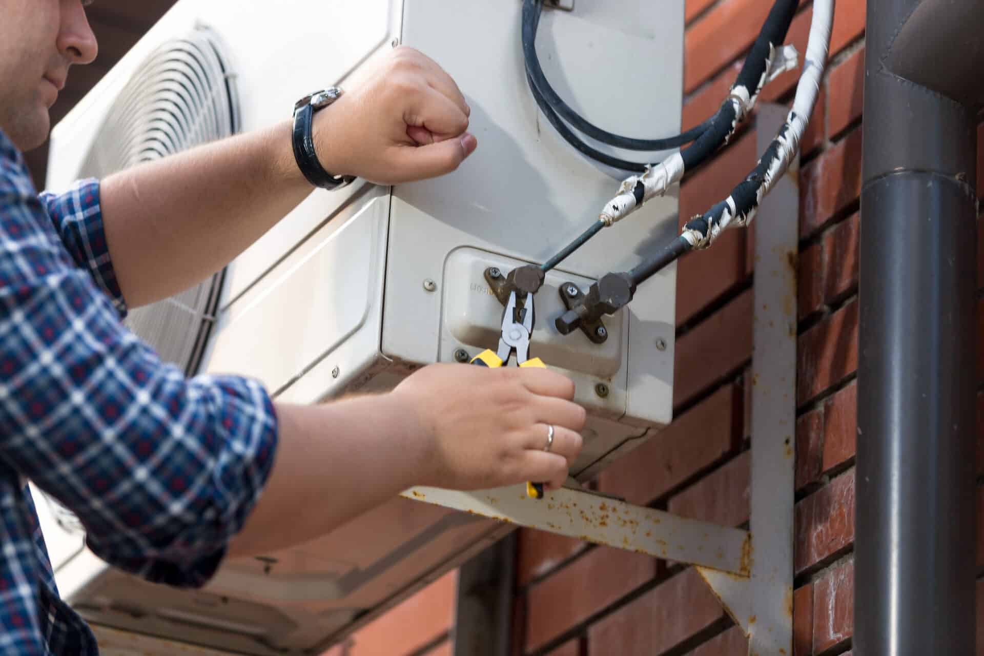 Technician using pliers on the pipe connections of a wall-mounted outdoor air-conditioning unit beside a brick wall.